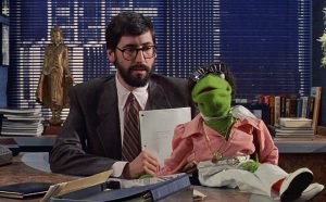 Kermit the frog dressed as a fast-talking agent sits on Leonard Winesop's desk. Leonard Winesop, played by John Landis, is a Broadway producer for the Winesop Theatrical Agency in The Muppets Take Manhattan. Targeted by Kermit (disguised as a boffo-socko agent), he listens to the frog's pitch for Manhattan Melodies, but refuses to even give it a read.
