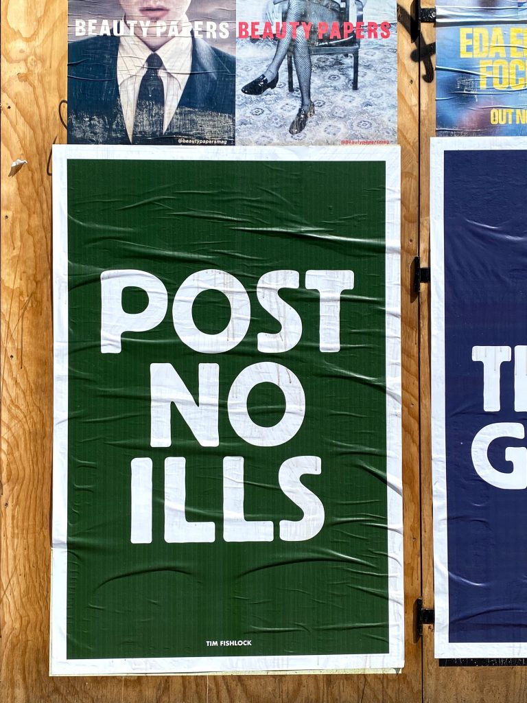 Plywood wall with various posters glued up featuring one that reads POST NO ILLS