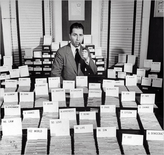Mortimer J. Adler holding a pipe in his left hand and mouth posing in front of dozens of boxes of index cards with topic headwords including 