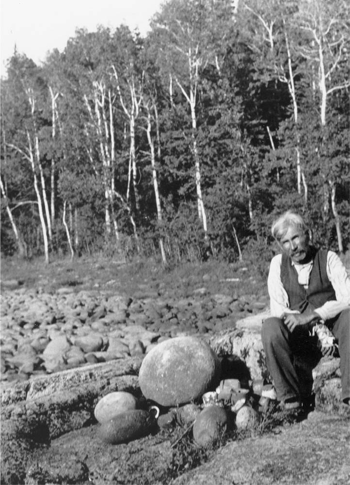 Black and white photo of a man in Western dress (pants, white shirt, and vest) sits on a rock with a forrest in the background. Beside him are several large round, but generally otherwise unremarkable rocks.