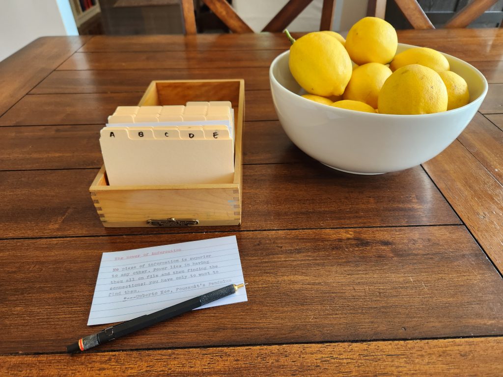 Angle down on a small, light brown wooden card index. The box has several manilla 1/5 cut 3x5" card dividers inside along with some white index cards. Outside of the box on the table in front of it are a typewritten index card and a black metal Rotring 800 0.5mm mechanical pencil. Off to one side is a white ceramic bowl full of lemons.