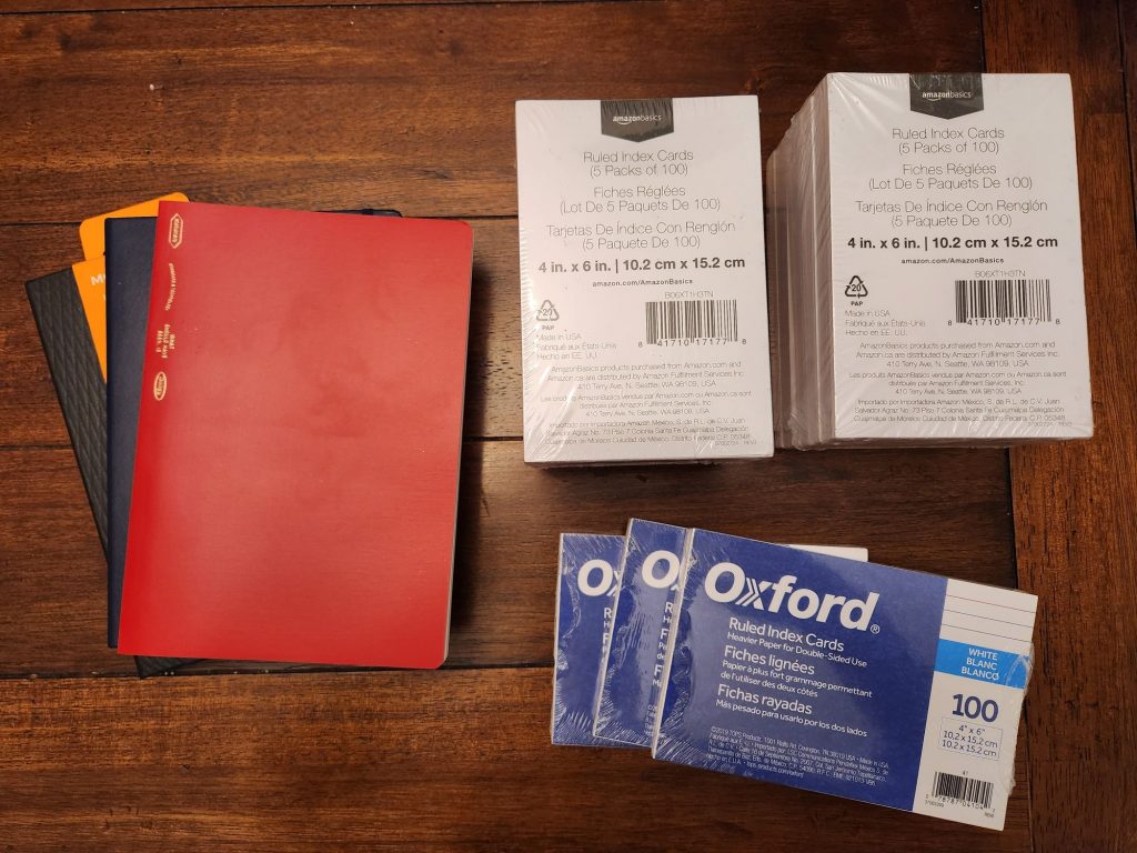 Three notebooks stacked up next to three separate piles of 1,300 index cards.