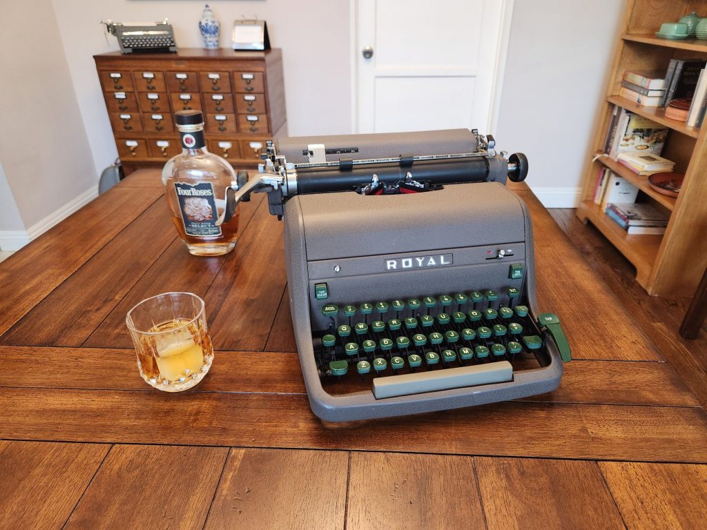A crystal old fashioned glass of bourbon on the rocks next to a Royal HH standard typewriter. In the background is a bottle of Four Roses bourbon and a 20 drawer library card catalog with another gray typewriter on it.