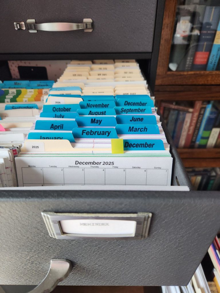 A Steelcase file index drawer open showing a four inch chunk of cards separated by tabbed cards featuring the names of the 12 months of the year and one divider in the back labeled 2026. On the front card is a calendar for the month of December 2025. In the blurry foreground is a label for the drawer of cards that reads "Memindex".