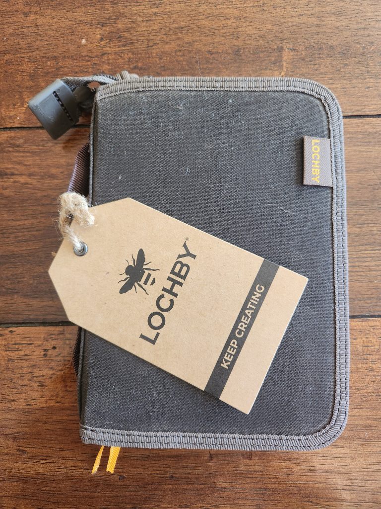 Lochby Field Folio A6 sits closed on a brown wooden table with it's Lochby kraft identification tag sitting on top of it. The exterior of the folio features brown waxed canvas.