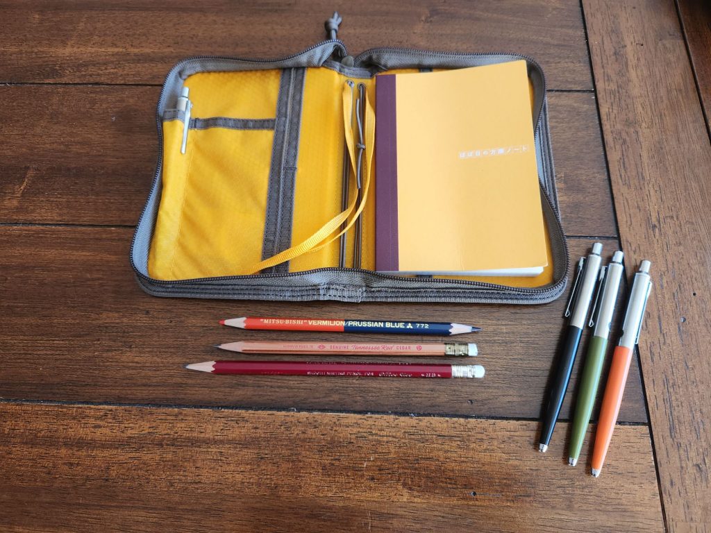 Lochby Field Folio A6 surrounded by several pens and pencils. Inside the right pocket of the folio is a yellow covered Hobonichi A6 notebook