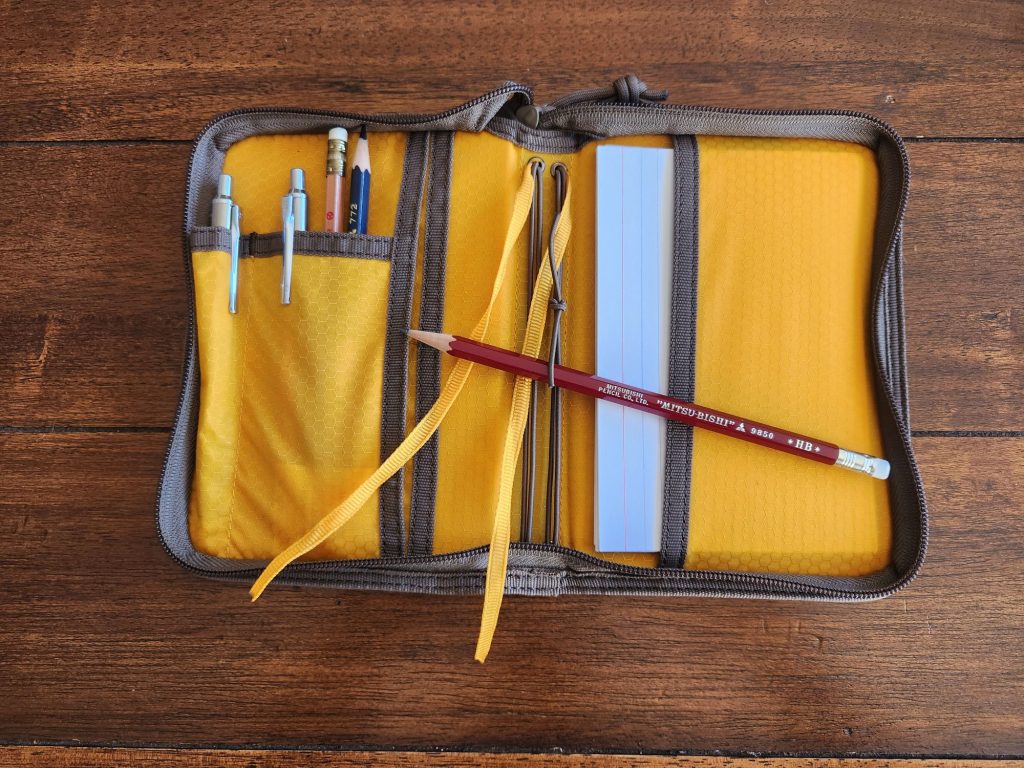 Lochby Field Folio A6 with a bunch of 4 x 6" index cards tucked into the right side interior pocket .The interior of the folio features yellow highlight material as well as two yellow ribbon bookmarks.  The left side has pocket space for several pens and pencils. Sitting crosswise across the top of the folio is a burgundy Mitsubishi 9850 HB pencil