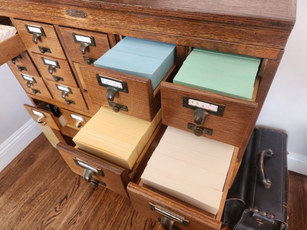 Four drawers of a library card catalog full of index cards in blue, green, buff, and ivory.