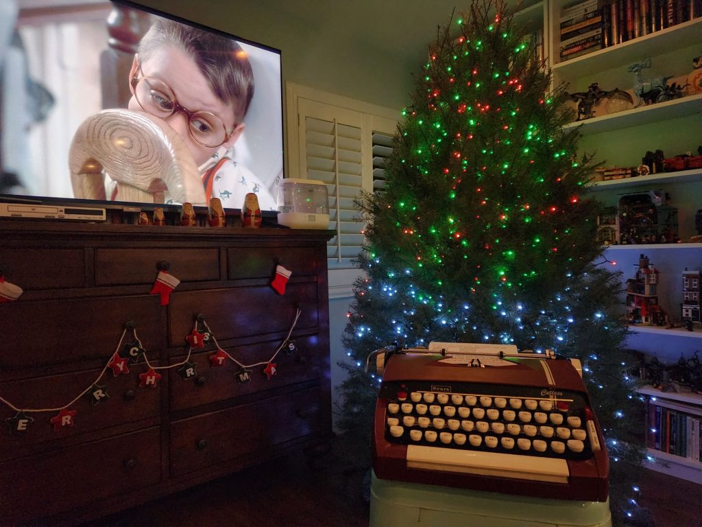 A 1964 Sears Cutlass sits on a stool in front of a Christmas tree. To the side is a wooden bureau with a wide screen television on it featuring the face of Kieran Culkin as Fuller in Home Alone. His face is being squished up against a chair and it gives the appearance that he's trying to look at the typewriter in front of the television.