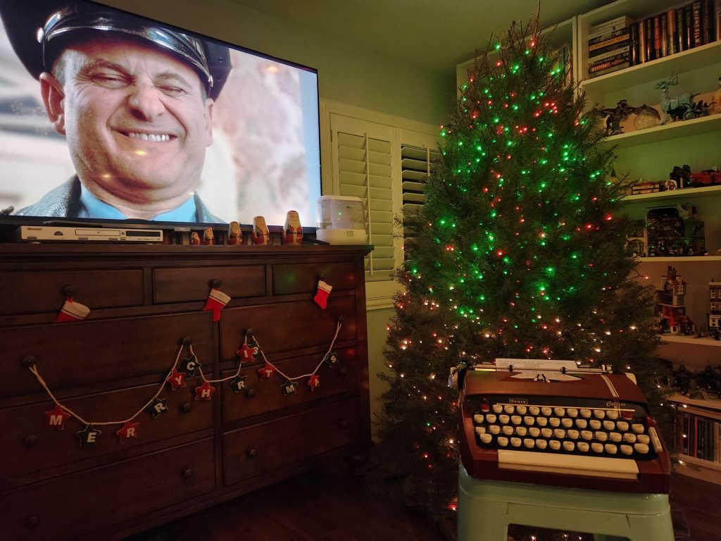 A 1964 Sears Cutlass sits on a stool in front of a Christmas tree. To the side is a wooden bureau with a wide screen television on it featuring the face of Joe Pesci as Harry in Home Alone. It almost appears as if he's squinting at the typewriter.
