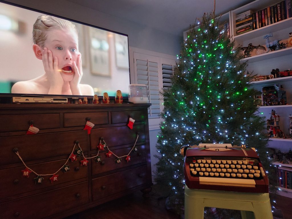 A 1964 Sears Cutlass sits on a stool in front of a Christmas tree. To the side is a wooden bureau with a wide screen television on it featuring the face of Macauley Culkin as Kevin in Home Alone as he claps his hands to his face and screams.