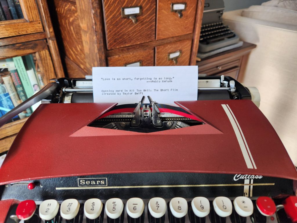 A burgundy 1964 Sears Cutlass typewriter with white racing strips which has a typed index card in the platen that reads: "Love is so short, forgetting is so long." ---Pablo Neruda | Opening card in All Too Well: The Short Film directed by Taylor Swift