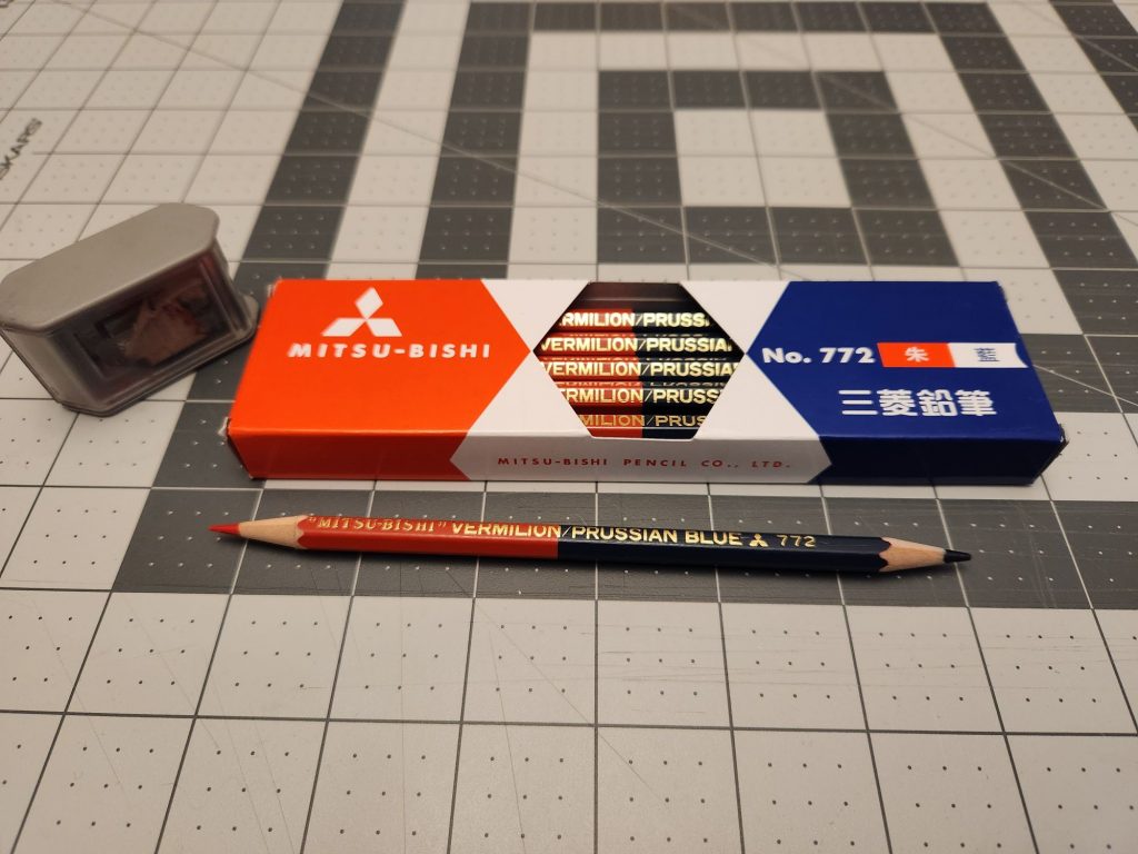 A box of Mitsubishi No. 772 pencils with a sharpened one sitting in front of it. One end of the half red/half blue pencil is vermillion and the other end is Prussian blue.