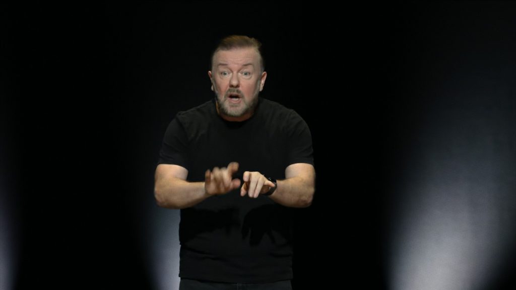 Ricky Gervis in a black t-shirt pretends to be typing as if at an invisible typewriter