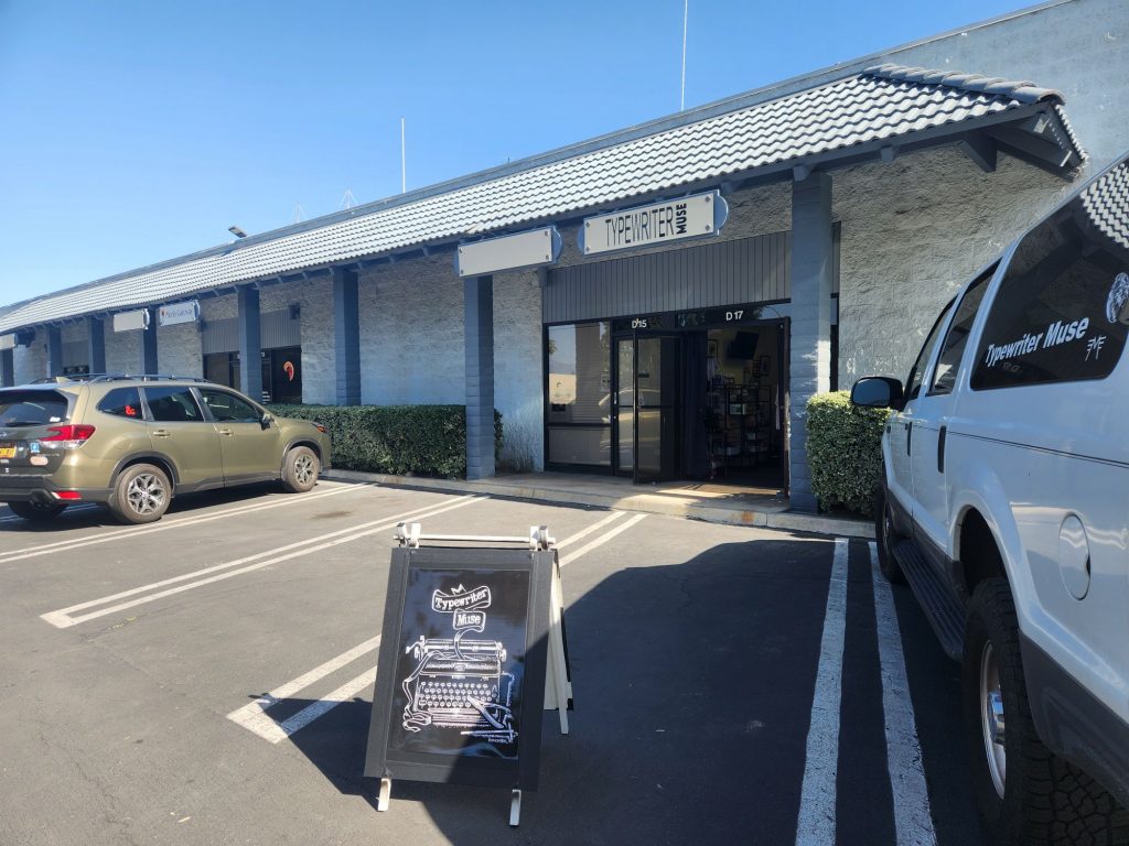 Single story business park building featuring the storefront of Typewriter Muse including a black sandwich board touting the store.