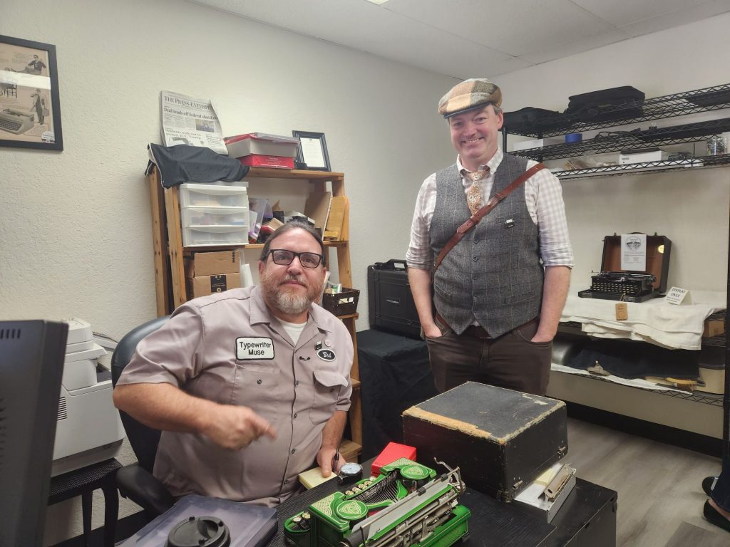 Bob Marshall with his hair tied back and wearing his mechanic's shirt sitting at the intake desk in front of a green Celtic-painted Royal P owned by Bryan Mahoney who is wearing a plaid vest and flat cap.