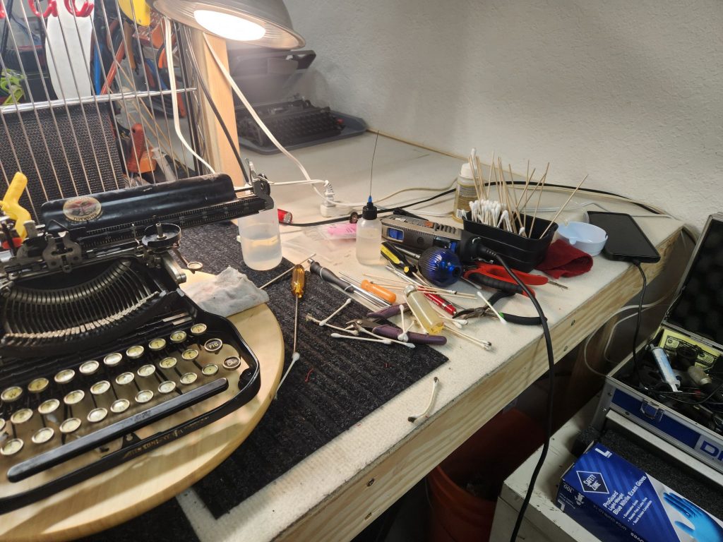 A typewriter mechanics's station with a Corona 3 on one side and a bunch of long handled q-tips  and other cleaning tools on the other side.
