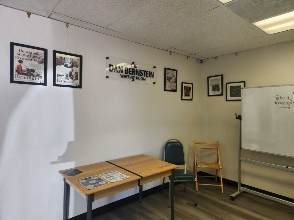 A view of the front reception/writing room at Typewriter Muse. There are several small desks with typewriters and a dedicatory sign on the wall that reads "The Dan Bernstein Writing Room".