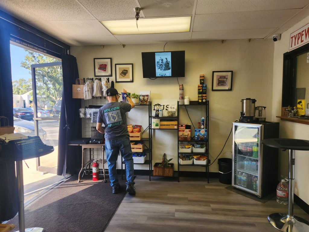 Front entrance at Typewriter Muse with a snack station and small drink refrigerator next to a typewriter table and a ribbon dispenser.