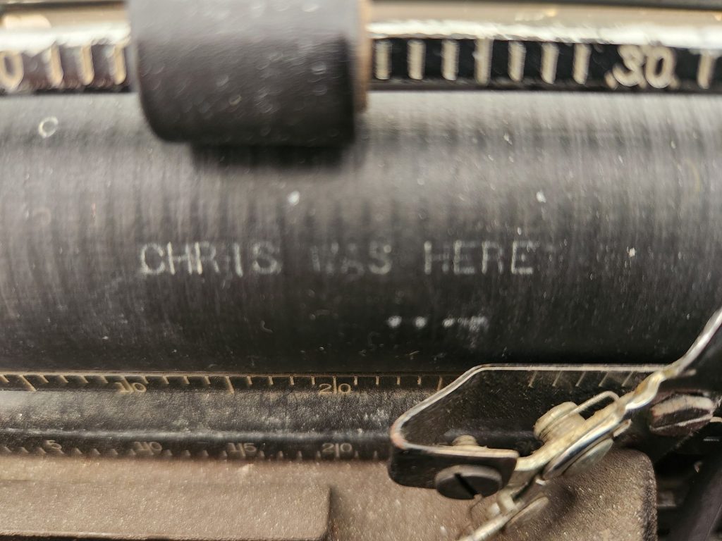 Capital white gothic letters typed onto a platen read: "CHRIS WAS HERE"