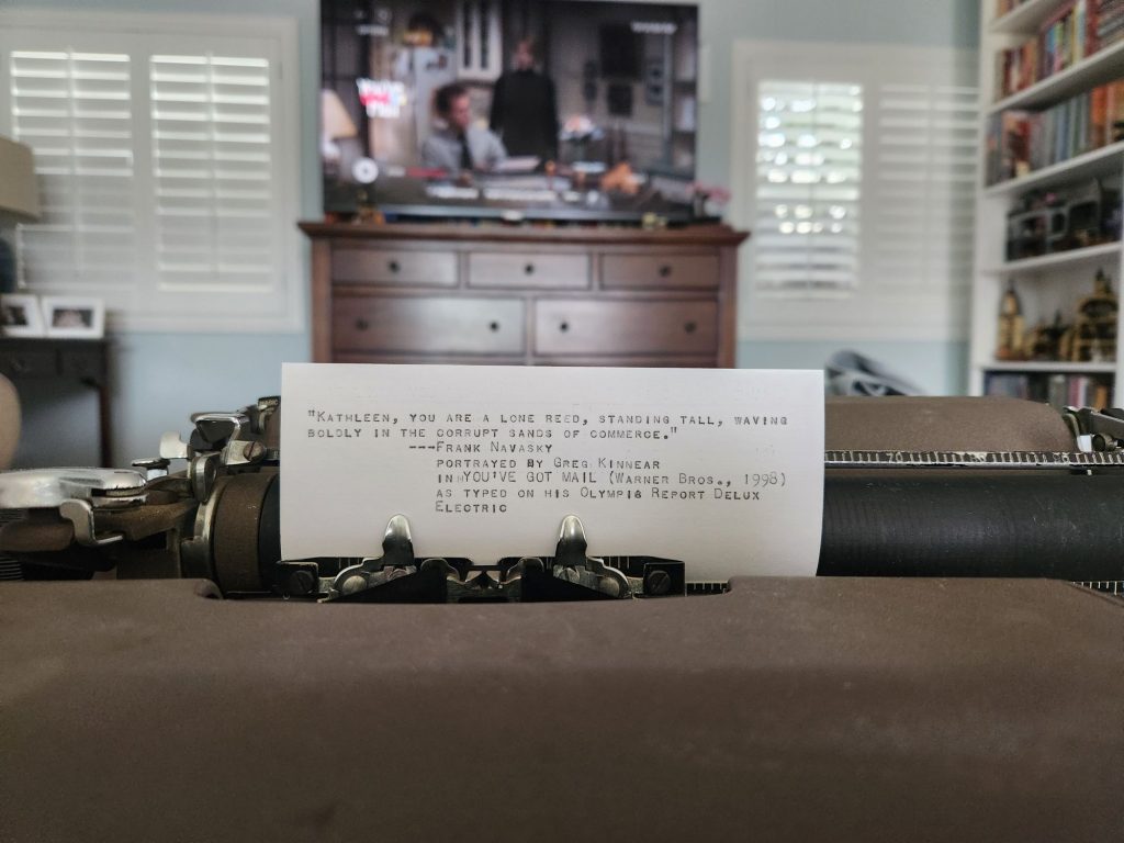 Close up of an index card in a typewriter carriage which reads: "Kathleen, you are a lone reed, standing tall, waving boldly in the corrupt sands of commerce." -Frank Navasky | You've Got Mail (Warner Bros., 1998) | as typed on his Olympia Report De Luxe Electric. In the background is a wooden dresser with a large screen television which has a somewhat blurry image of Greg Kinnear sitting at his typewriter from the movie.