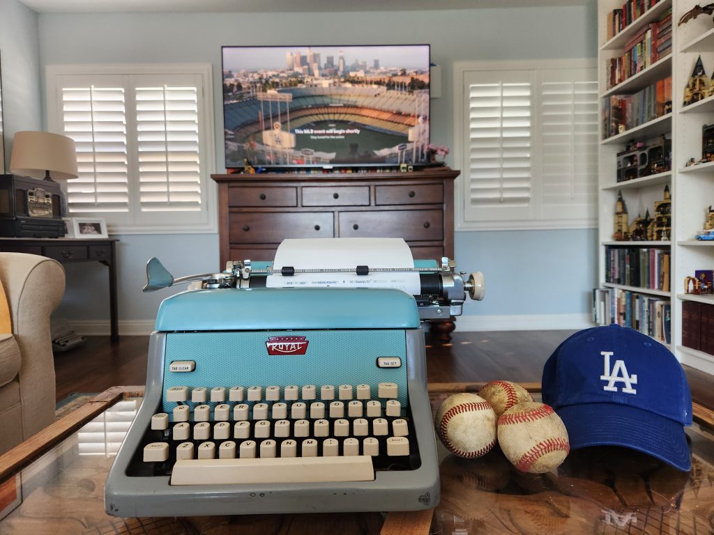 "Dodger Blue" 1957 Royal FP typewriter at the ready in front of a television featuring Dodger Stadium to score the baseball game. Next to the typewriter on its table are three baseballs and a blue Dodgers cap featuring the iconic LA logo.