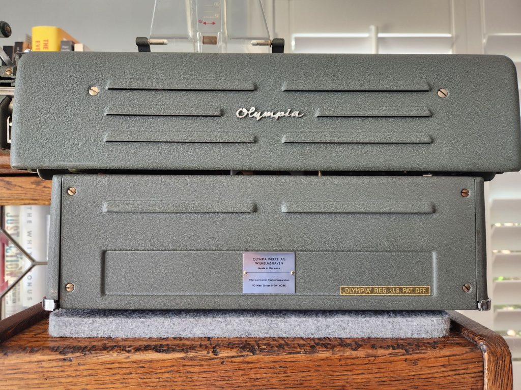 Rear of a green crinkle painted Olympia SG1.  The bottom of the machine has a panel with a metal Olympia manufacturer's plate showing the company name, place of manufacture, and location of the dealer ship in New York. On either side is a wide rectangle where other identifiers could be similarly placed.