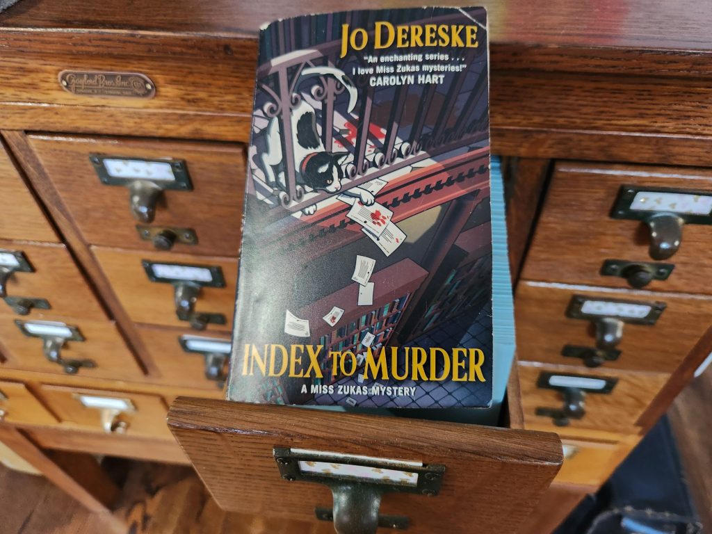 Paperback book Index to Murder by Jo Dereske sitting on a library card catalog. The cover of the book features a cat on the second story of a library playing with some index cards with bloody paw prints and knocking them past a railing to the floor below.