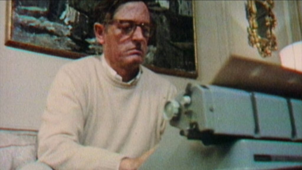 William F. Buckley in a white shirt with a sweater over it and wearing his signature glasses begins a new line of writing on a Hermes 3000 typewriter