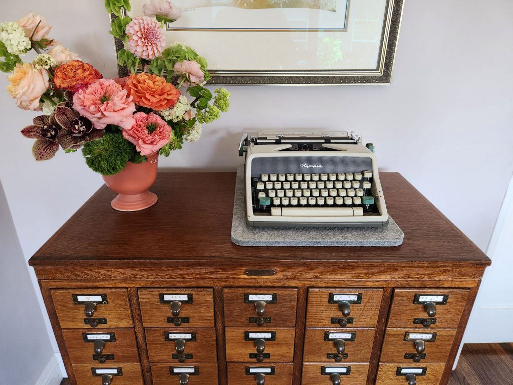 Front view of a 1962 Olympia SM7 typewriter sitting on a library card catalog next to a vase of flowers.