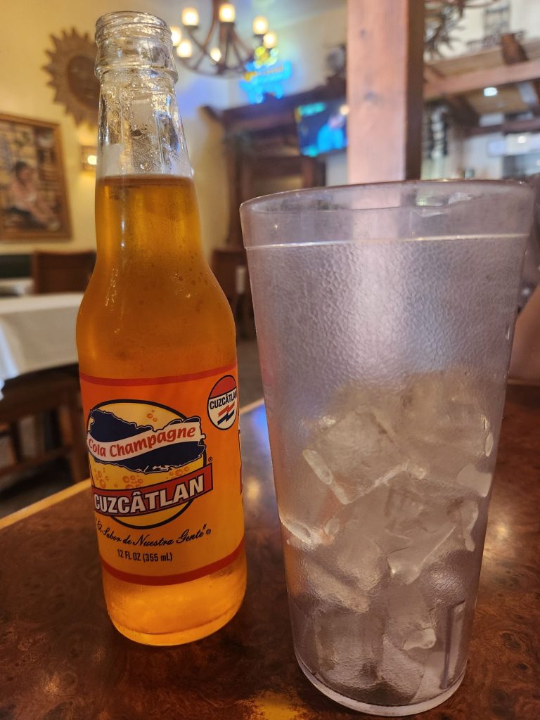A glass bottle of Cuzcatlan brand cola champagne in a lovely orange color next to a glass of ice at a resturant