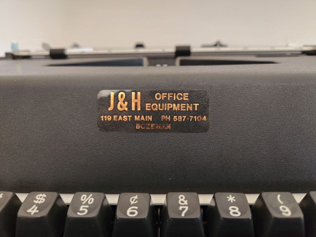 Close up of a sales and service sticker on the hood of a Olympia Report de Luxe typewriter. In gray with gold lettering it reads "J&H Office Equipment, 119 E. Main | PH 587-7104 | Bozeman