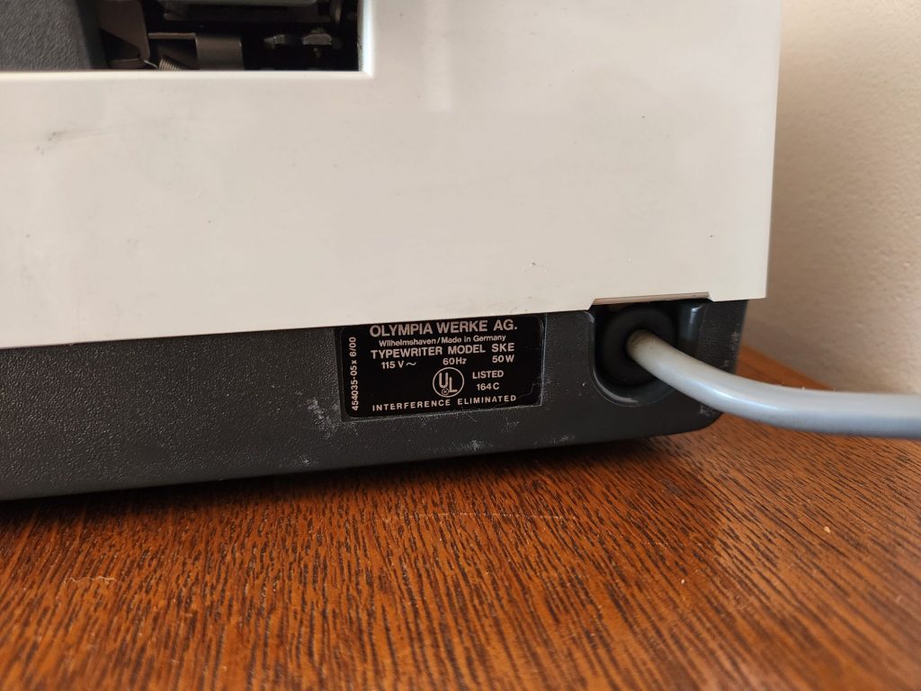 Close up of a model sticker on the lower right side of a Olympia Report de Luxe typewriter next to it's power cord. We see that it's listed as "typewriter model SKE" and that it uses 115V, 60Hz, 50 Watt power and is UL listed.