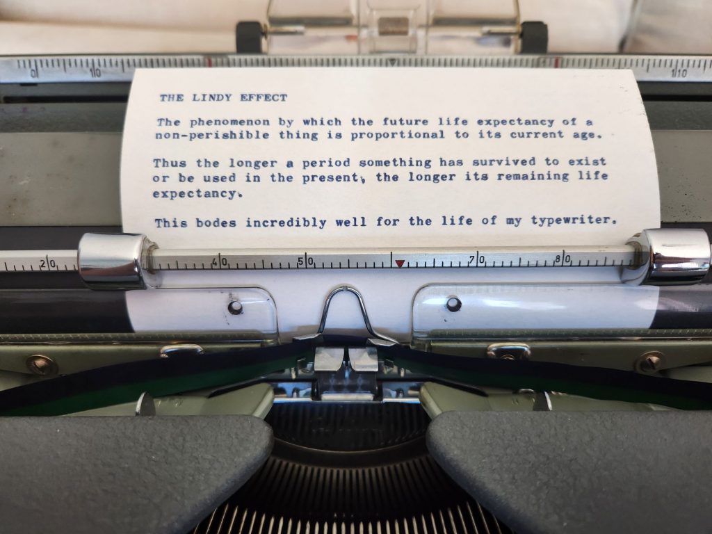 A typed index card in the platen of a typewriter which reads: "The Lindy Effect | The phenomenon by which the future life expectancy of a non-perishable thing is proportional to its current age.  | Thus the longer a period something has survived to exist or be used in the present, the ;onger its remaining life expectancy. | This bodes incredibly well for the life of my typewriter."