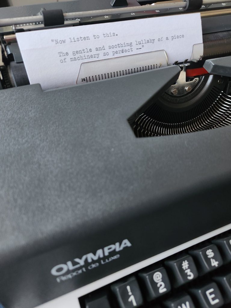 A 1977 Olympia Report de Luxe electric typebar typewriter with an index card in the carriage that reads: "Now listen to this. The gentle and soothing lullaby of a piece of machinery so perfect--"