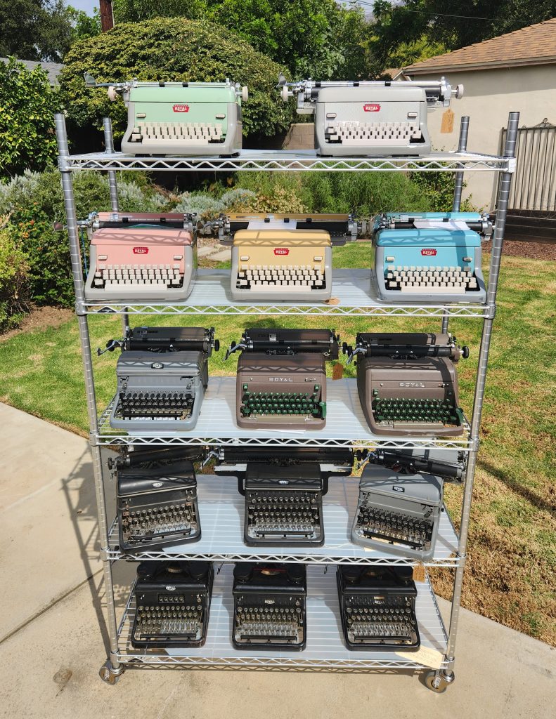 A wire metal shelving unite on concrete in front of a grassy yard with some bushes and flowers in the background. On the 5 shelves are a collection of Royal Standard typewriters with three on each shelf except the top which only has two.