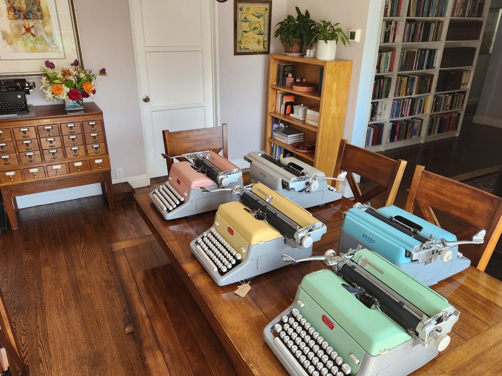 A dining room table with two rows of Royal FP standard typewriters set up for use. In the background are two different bookcases and a library card catalog with a flower arrangement next to a black typewriter