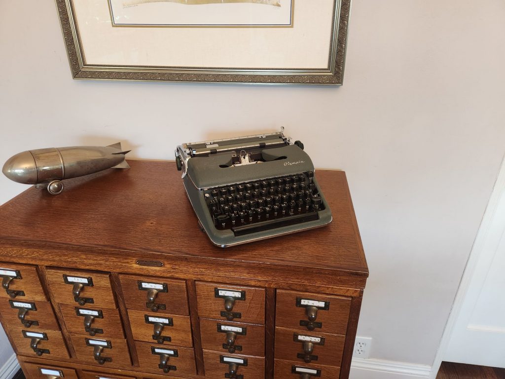 1959 Olympia SM3 portable typewriter in green crinkle paint on an oak library card catalog next to a silver plated cocktail shaker in the shape of a zeppelin.