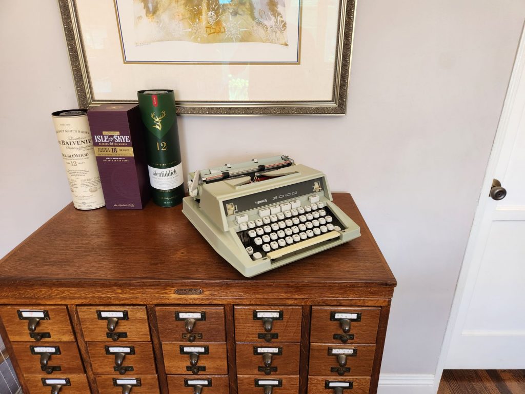 A greenish gray 1971 Hermes 3000 with a boxy plastic body sits on a library card catalog next to three gift bottles of Scotch.