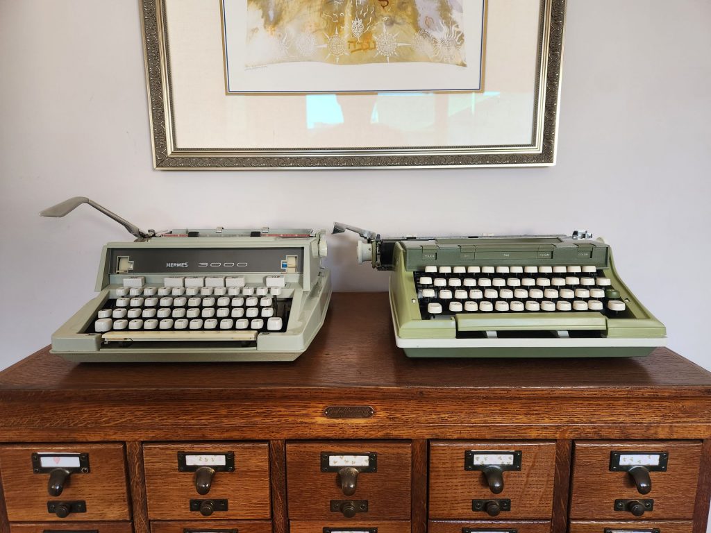 A light green 1971 boxy Hermes 3000 with greyish-cream keys sits to the right of a 1971 avocado green Sears Newport typewriter