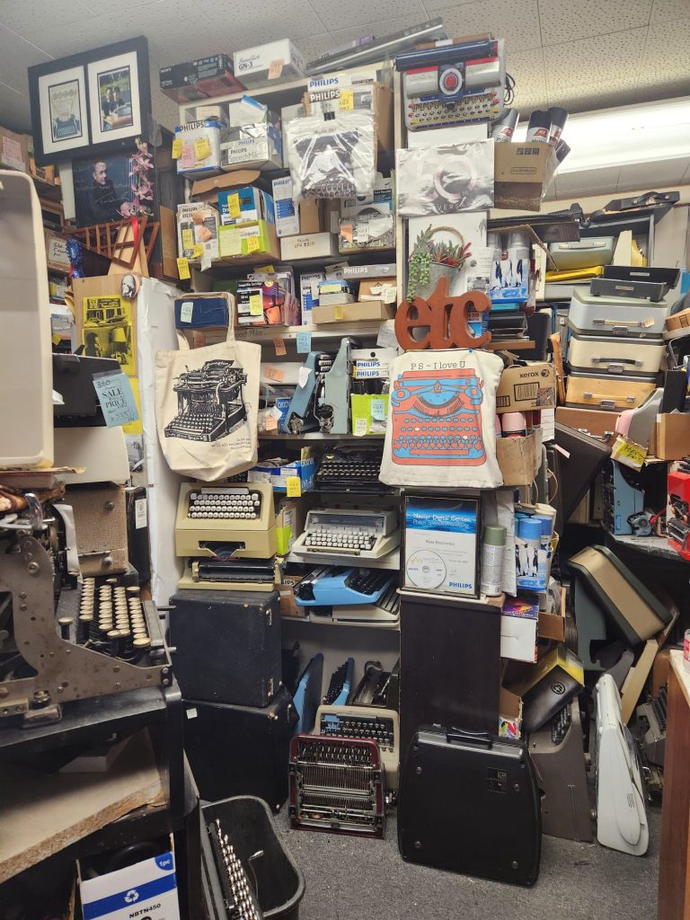 A bookcase crammed floor to ceiling with typewriters, and decorative items including book bags with typewriters printed on them.